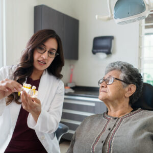 A latin female dentist and a senior patient looking at model teeth in the dental room.
