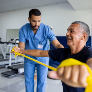 Mature Latin American man doing physical therapy exercises using a stretch band with the assistance of his therapist