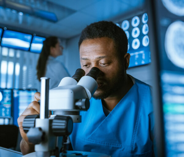 Medical Research Scientist Looking under the Microscope in the Laboratory. Neurologist Solving Puzzles of the Mind and Brain. In the Laboratory with Multiple Screens Showing MRI / CT Brain Scan Images.